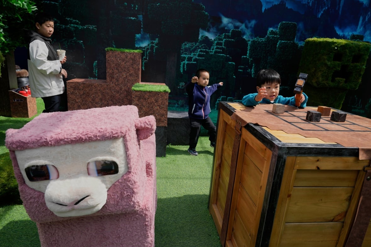 Three children interact with a display of green grass, a pink square sheep, and a hammer on a table